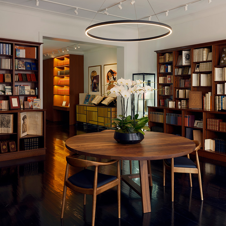 A cosy library with dark wood floors, shelves brimming like the best London bookshops, a round wooden table with four chairs, a potted orchid centrepiece, and a modern circular light above. Artworks and display shelves complete the inviting scene.