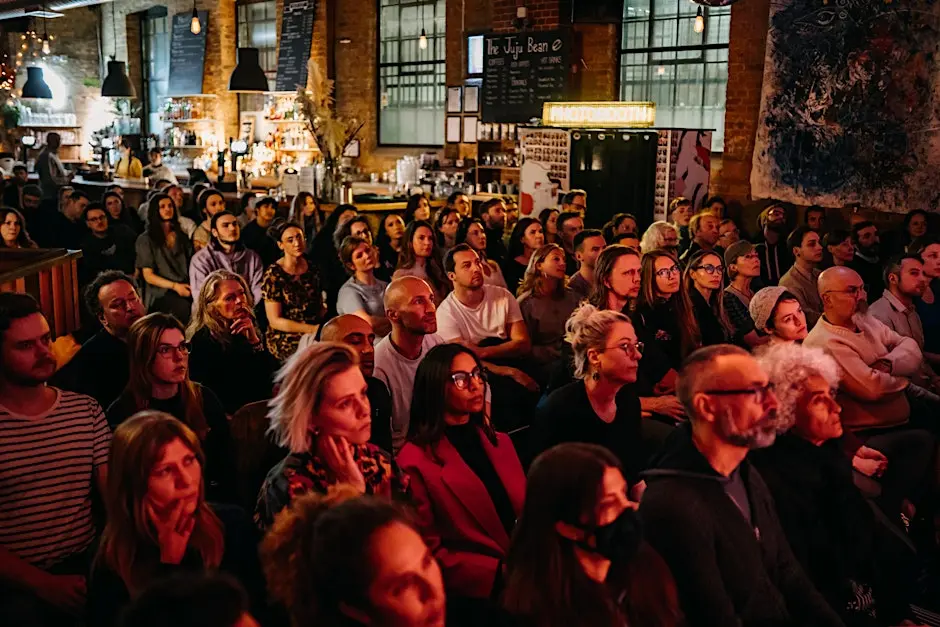 A diverse group of people sits closely together in a warmly lit indoor venue, attentively watching an unseen pub lecture. The background features industrial-style decor, large windows, and a menu board.