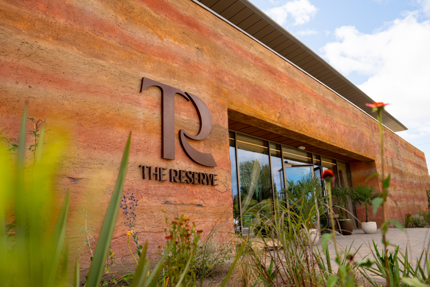 A modern building with a textured, earth-toned façade features a large metal logo and THE RESERVE sign by the entrance, hinting at exclusive zoo stays. Tall plants and flowers are in the foreground under a partly cloudy sky.