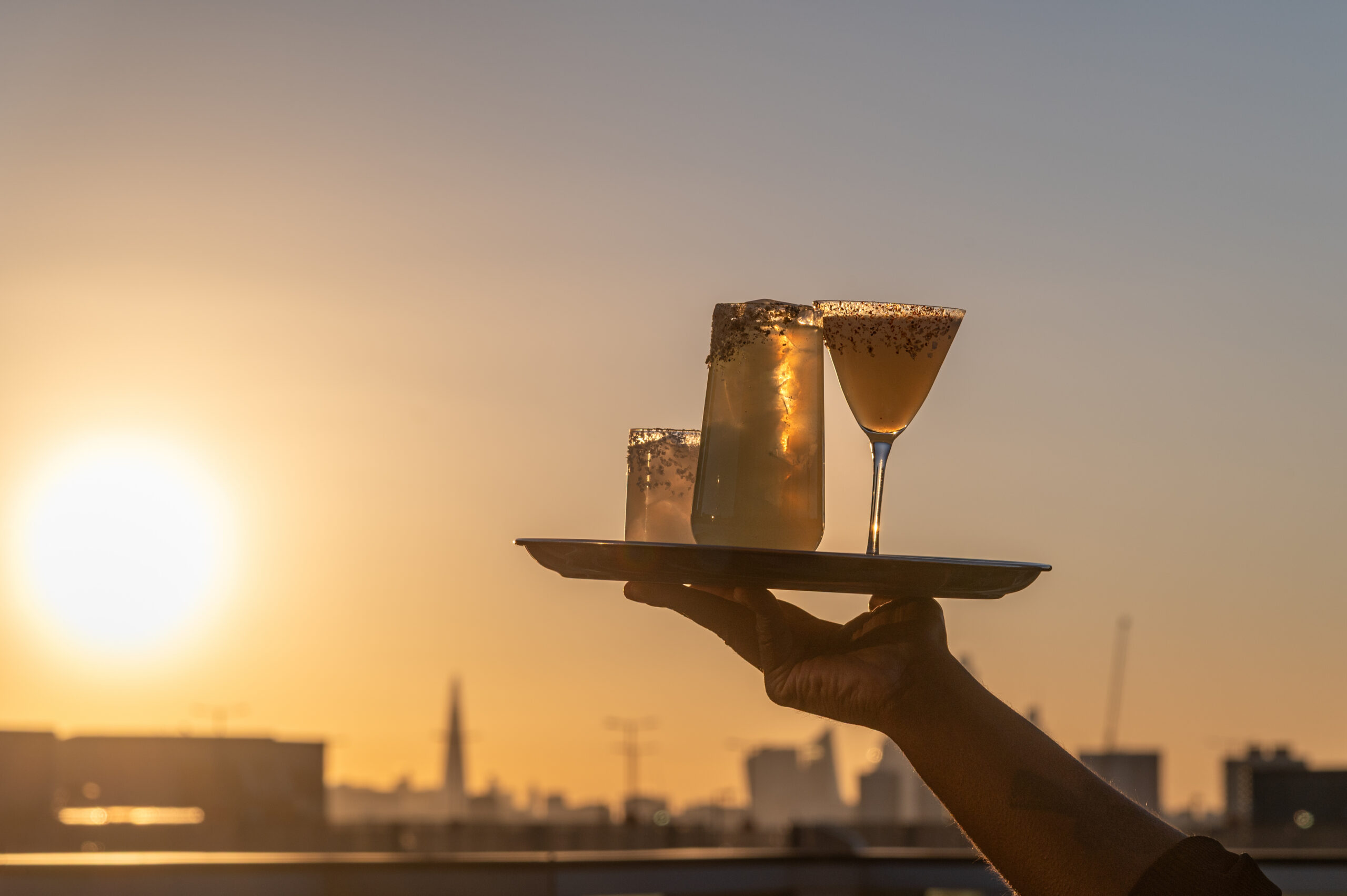 A hand holds a tray with three cocktails garnished with sugar rims, raised against a city skyline at sunset—perfect for enjoying along the margarita mile on a bright, clear evening.
