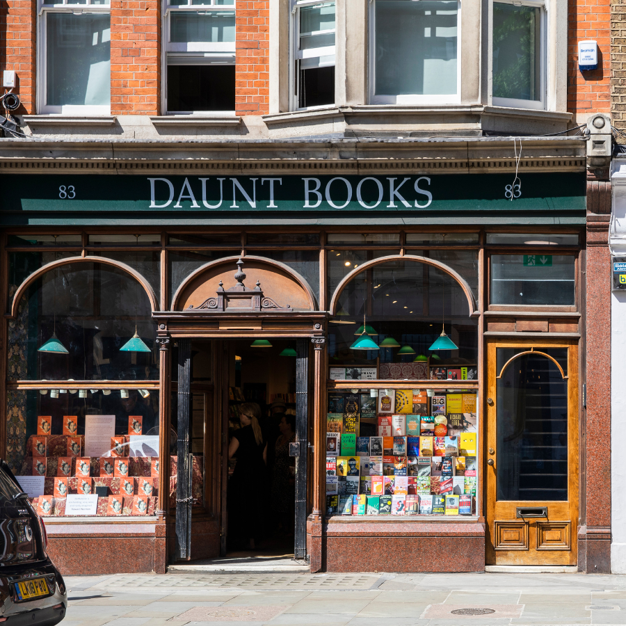 The exterior of Daunt Books, one of the best London bookshops, features a green sign, arched doorway, and large windows displaying colourful books and covers, all set in a brick building on a sunny day.