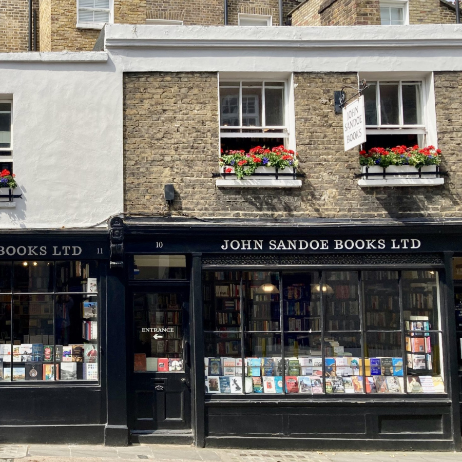 A charming bookshop with large front windows filled with books, black trim, and flower boxes under the upper windows. John Sandoe Books Ltd., set in a historic brick building, is renowned as one of the best London bookshops.