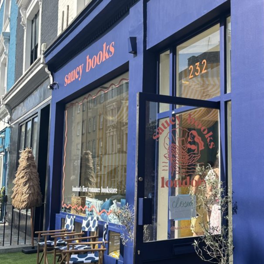 A blue shopfront with red lettering reading “saucy books,” advertising itself as “London’s first romance bookshop.” The door is open, with two patterned chairs and plants outside—one of the best London bookshops for romance lovers.