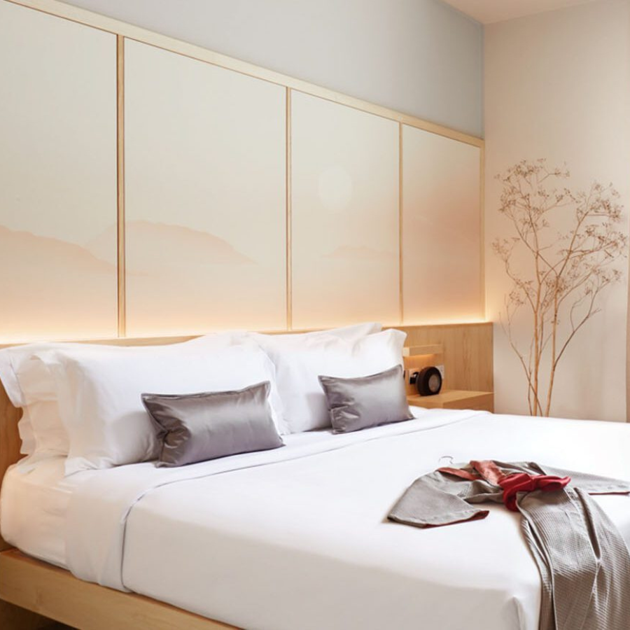 A neatly made bed with white linens and grey accent pillows in a modern, minimal bedroom—perfect inspiration for your weekend guide. Soft lighting highlights a panelled headboard, while a delicate dried plant stands elegantly in the corner.