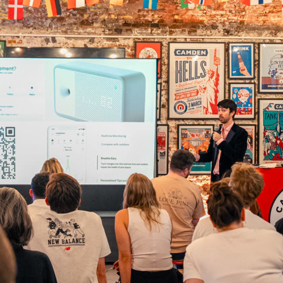 A man gives a pub lecture-style presentation to an audience in a colourful, poster-filled room. He stands beside a large screen displaying product images, QR codes, and graphs whilst international flags hang above the crowd.