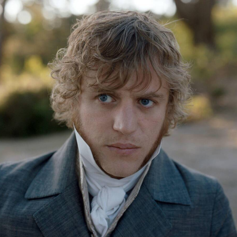 A young man with curly blond hair and blue eyes, evoking classic romantic heroes like Gatsby or Romeo, wears early 19th-century clothing—a high-collared shirt, cravat, and grey coat—as he stands outdoors with a serious expression.