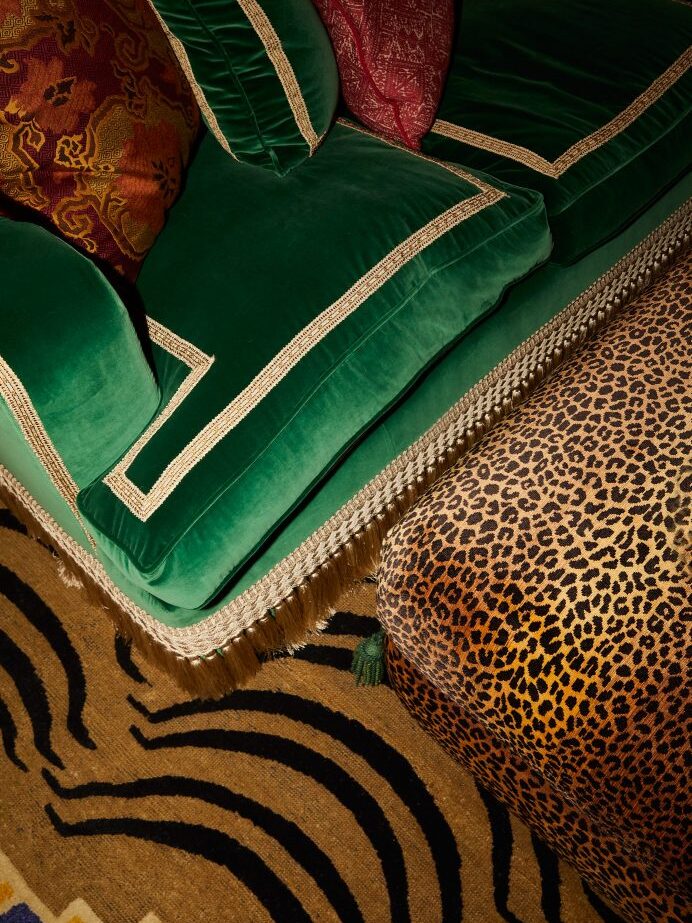 A green velvet sofa with gold trim and red cushions, inspired by Barlow & Barlow, sits next to a footstool upholstered in leopard print fabric, all on a patterned brown and black rug.