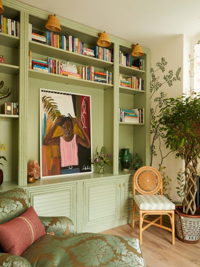 Cosy reading nook with sage green built-in bookshelves, books, and rattan wall lamps. Interiors Insider highlights a large portrait painting on a lower shelf, a patterned chaise longue, rattan chair, and potted plants nearby.
