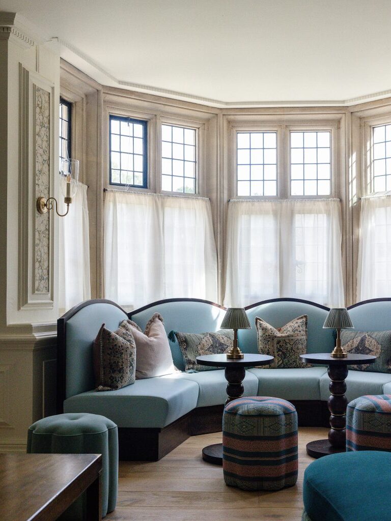 A bright, elegant sitting area by Barlow & Barlow features curved light blue sofas, patterned cushions, round wooden tables, matching stools, and tall bay windows with sheer white curtains letting in natural light.