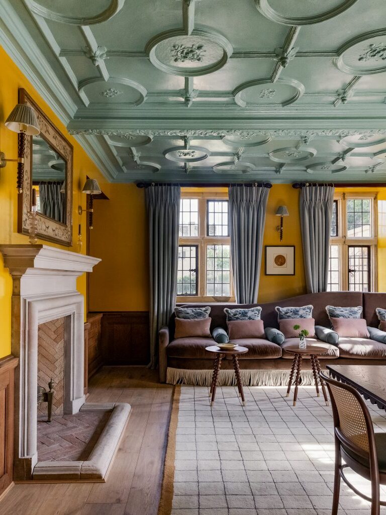 A traditional living room by Barlow & Barlow features a decorative green coffered ceiling, yellow walls, tall windows with blue curtains, a white fireplace, a patterned rug, and a long sofa with cushions and small wooden tables.