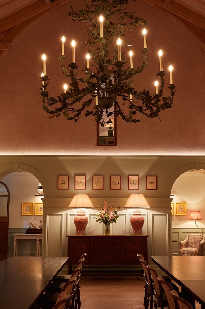 A warmly lit dining room by Barlow & Barlow features a large leafy chandelier with candles, a wooden table and chairs, a sideboard with two lamps, framed art on the wall, and arched doorways leading to softly lit adjoining rooms.