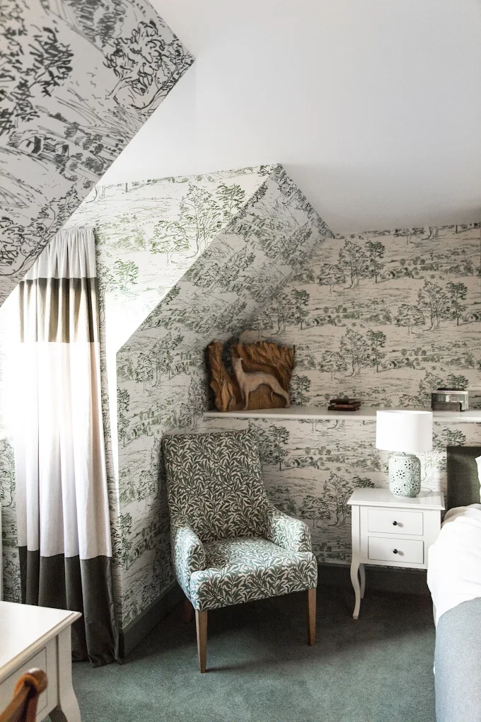 A cosy bedroom corner inspired by Kinloch Lodge, featuring nature-themed wallpaper, an upholstered armchair with leafy patterns, a white bedside table and lamp, and a wooden dog sculpture on a shelf under the sloped ceiling. Light filters through a window.