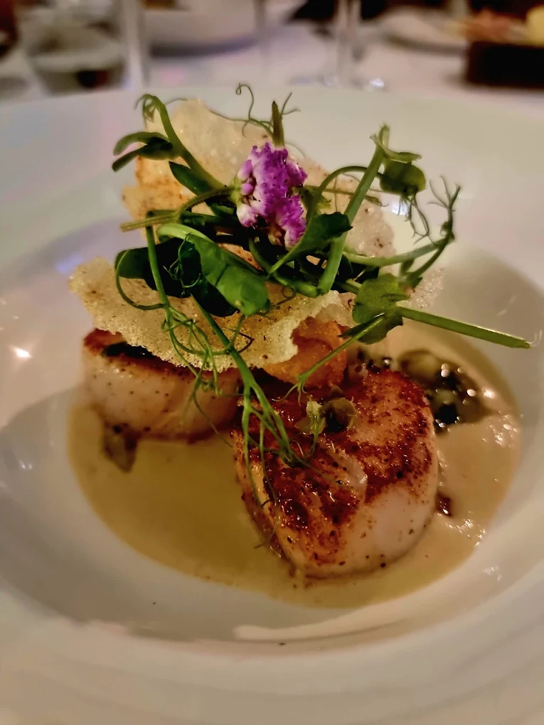Two seared scallops are topped with crispy garnish, microgreens, and an edible flower, served on a creamy sauce in a white bowl—a refined presentation inspired by Kinloch Lodge, with vibrant greens and purple accents.