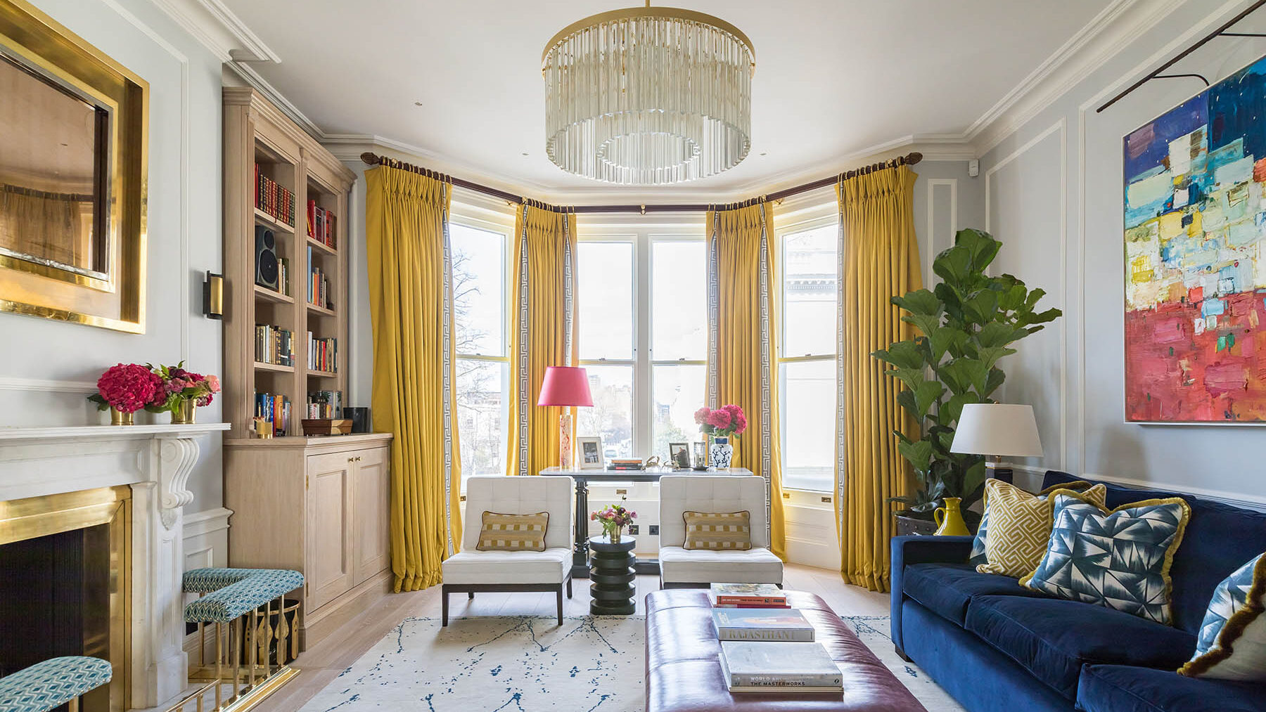 Bright living room with large windows, yellow curtains, a blue velvet sofa, colourful abstract art, a leather ottoman, and a fireplace with a gold-framed mirror—this Barlow & Barlow design exudes contemporary style and Interiors Insider flair.