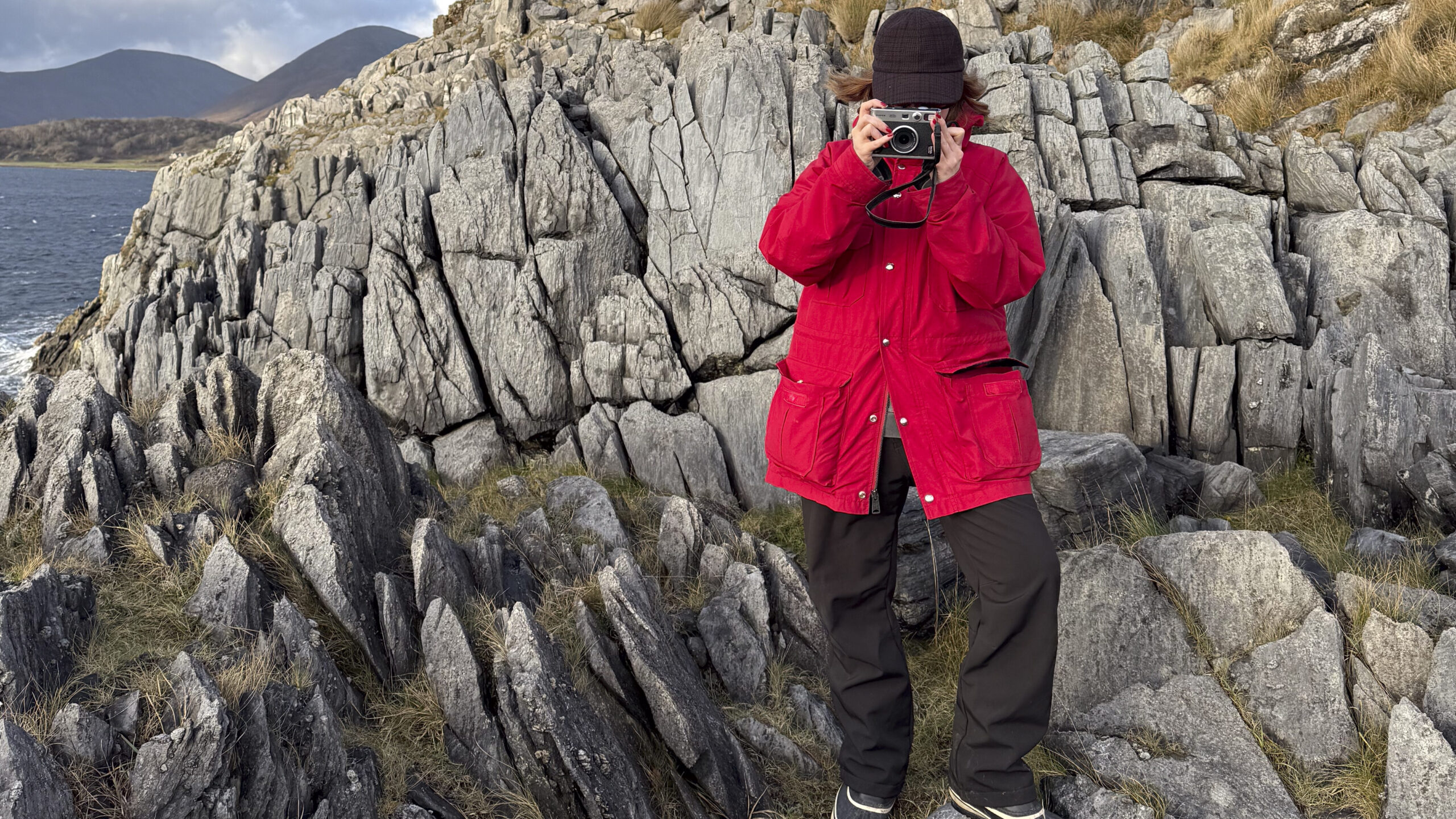 A person wearing a red jacket and brown hat stands on rocky terrain near Kinloch Lodge, holding a camera up to their face. Rugged grey rocks and grassy patches surround them, with hills and water visible in the background.