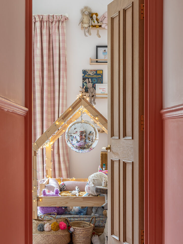 A cosy children’s room by Barlow & Barlow, with pink curtains, a wooden playhouse bed adorned with fairy lights and a disco ball, plus baskets of toys and wool—all glimpsed through an open wooden door. Featured on Interiors Insider.