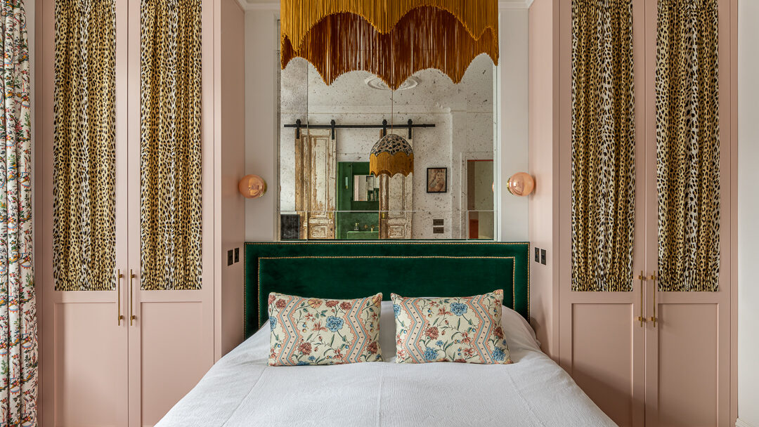 A stylish bedroom by Barlow & Barlow features a green velvet headboard, floral pillows, pink wardrobes with leopard print panels, gold light fittings, and a gold fringe pendant lamp, all reflected in a large mirror behind the bed.