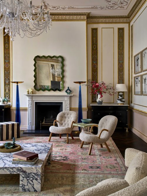 A luxurious living room by Barlow & Barlow with ornate gold-trimmed walls, a marble coffee table, plush armchairs, a decorative fireplace, crystal chandelier, and pastel vintage rug. Artworks and vases adorn this elegant Interiors Insider space.