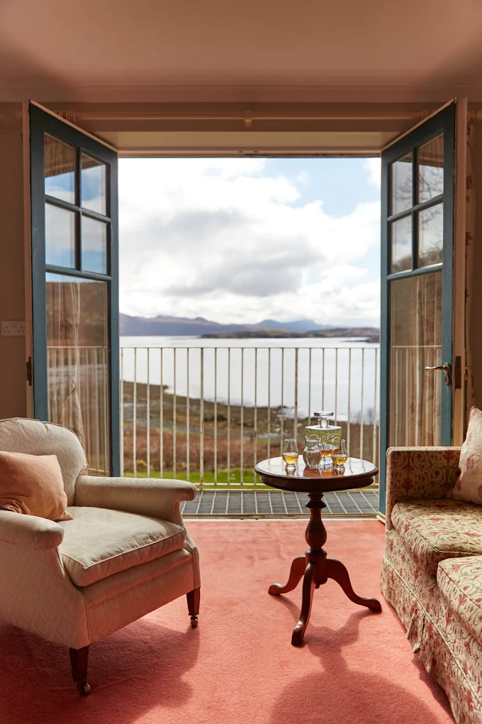A cosy sitting room at Kinloch Lodge features armchairs and a small round table with drinks on a pink carpet. Double doors open to a balcony overlooking a scenic lake and hills under a partly cloudy sky.