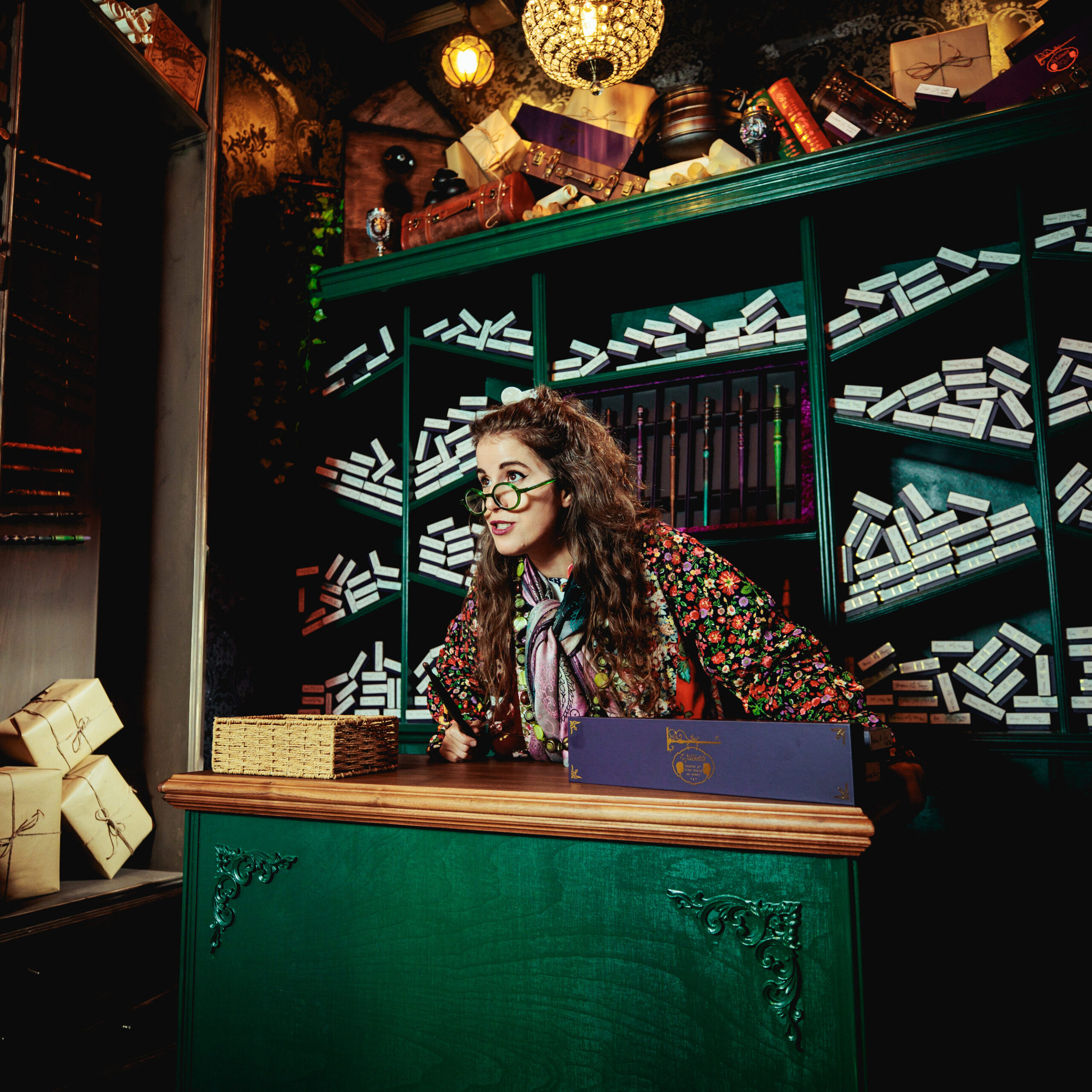 A woman with long curly hair, glasses, and colourful clothing leans excitedly over a green counter in a magical-looking shop inspired by Harry Potter bar restaurants and experiences London, filled with shelves of wand boxes and stacks of brown-wrapped parcels.