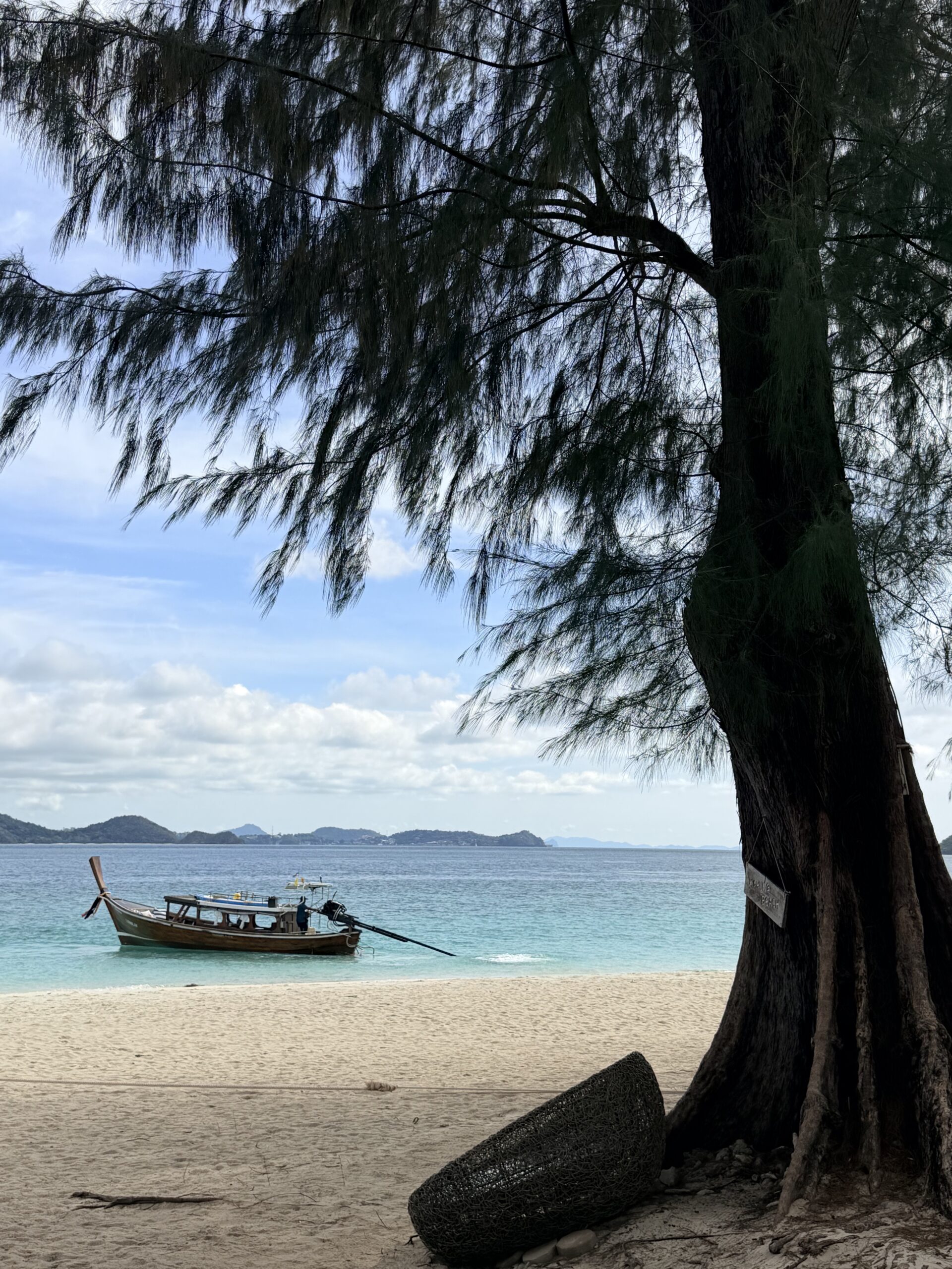 Traditional longtail boat anchored on a serene tropical beach framed by tall trees and crystal-clear blue water.