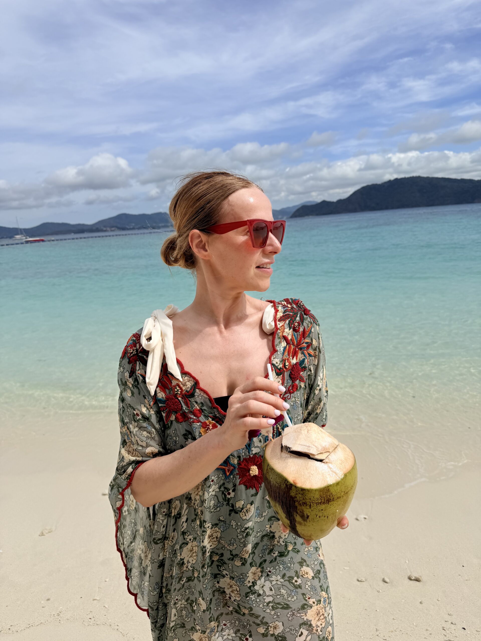 Woman in a floral beach cover-up holding a fresh coconut on a white sand beach with turquoise water and tropical island scenery.