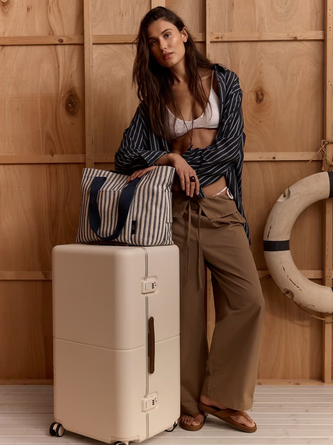 A woman in a striped shirt, white top, and brown wide-leg trousers leans on a cream-coloured suitcase from one of the best suitcase brands. A striped tote bag sits on top; she stands on wooden flooring, with a lifebuoy and wood-panelled wall behind her.