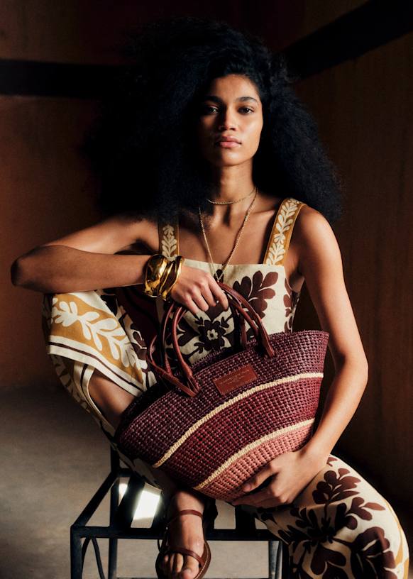A woman with voluminous curly hair sits on a chair, wearing a floral-patterned brown and cream jumpsuit. She holds one of this season’s chic basket bags and accessorises with gold jewellery and sandals in a softly lit room.