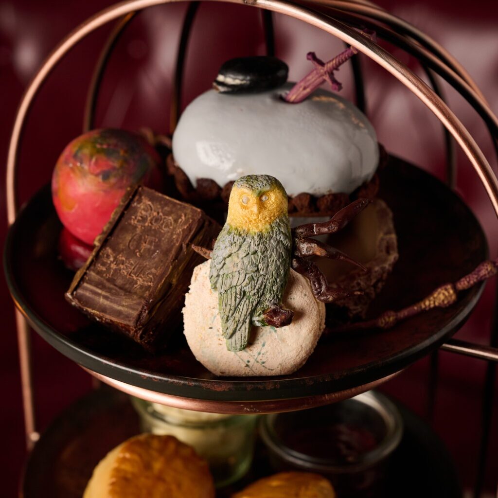 An assortment of desserts on a tiered tray, inspired by Harry Potter bar restaurants and experiences in London, featuring a white macaron topped with an owl figure, a glossy dome-shaped pastry, a decorated chocolate block, and a colourful confection.