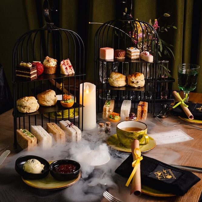 An elegant afternoon tea set-up inspired by Harry Potter bar restaurants and experiences in London, with tiered black cages holding assorted pastries, scones, and finger sandwiches. A teacup, plates, candle, and dry ice mist complete the magical table setting.