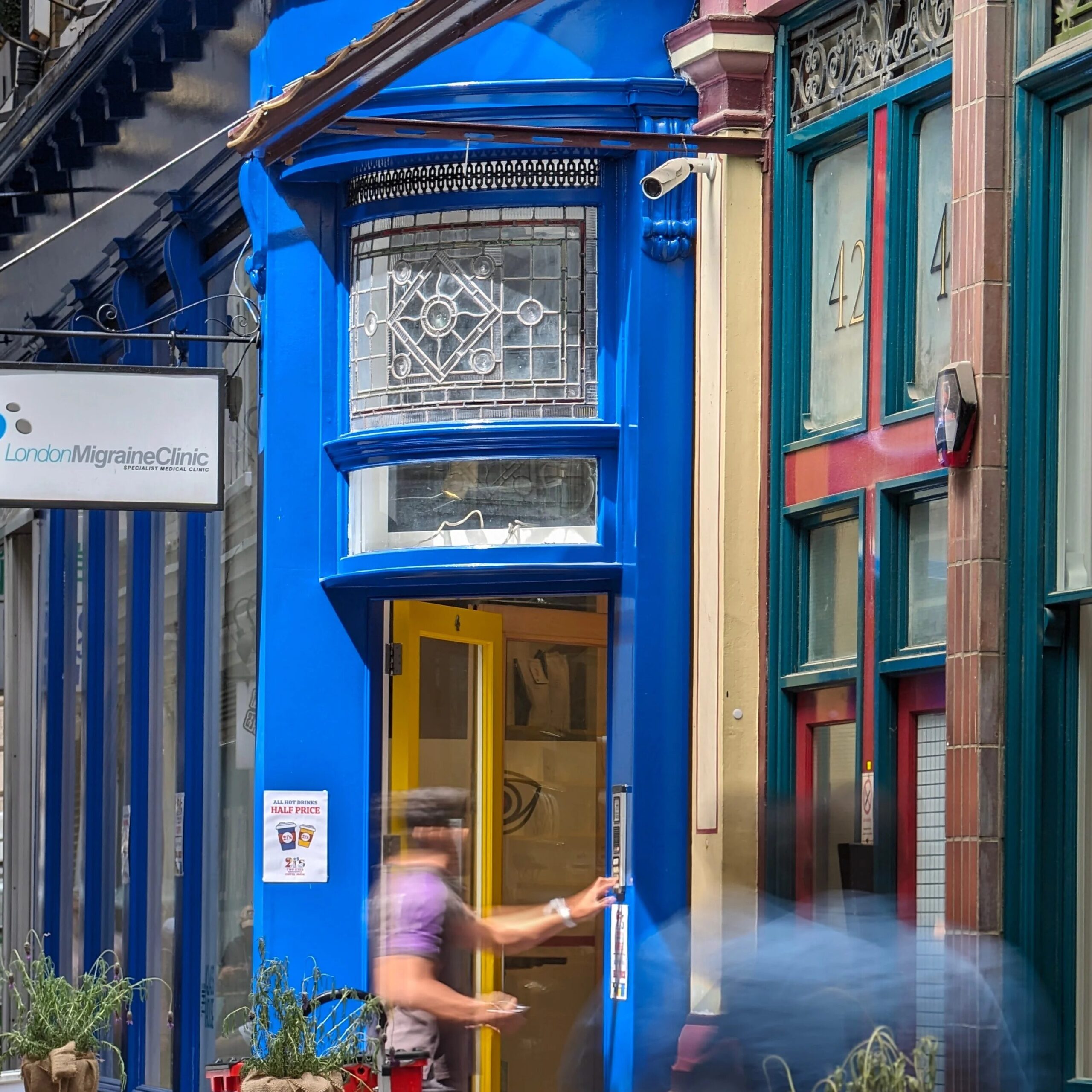 A person enters a vibrant blue shopfront with a yellow door, next to a sign reading London Migraine Clinic. The neighbouring building, numbered 42, hints at Harry Potter bar restaurants and experiences in London. The image is slightly blurred by motion.