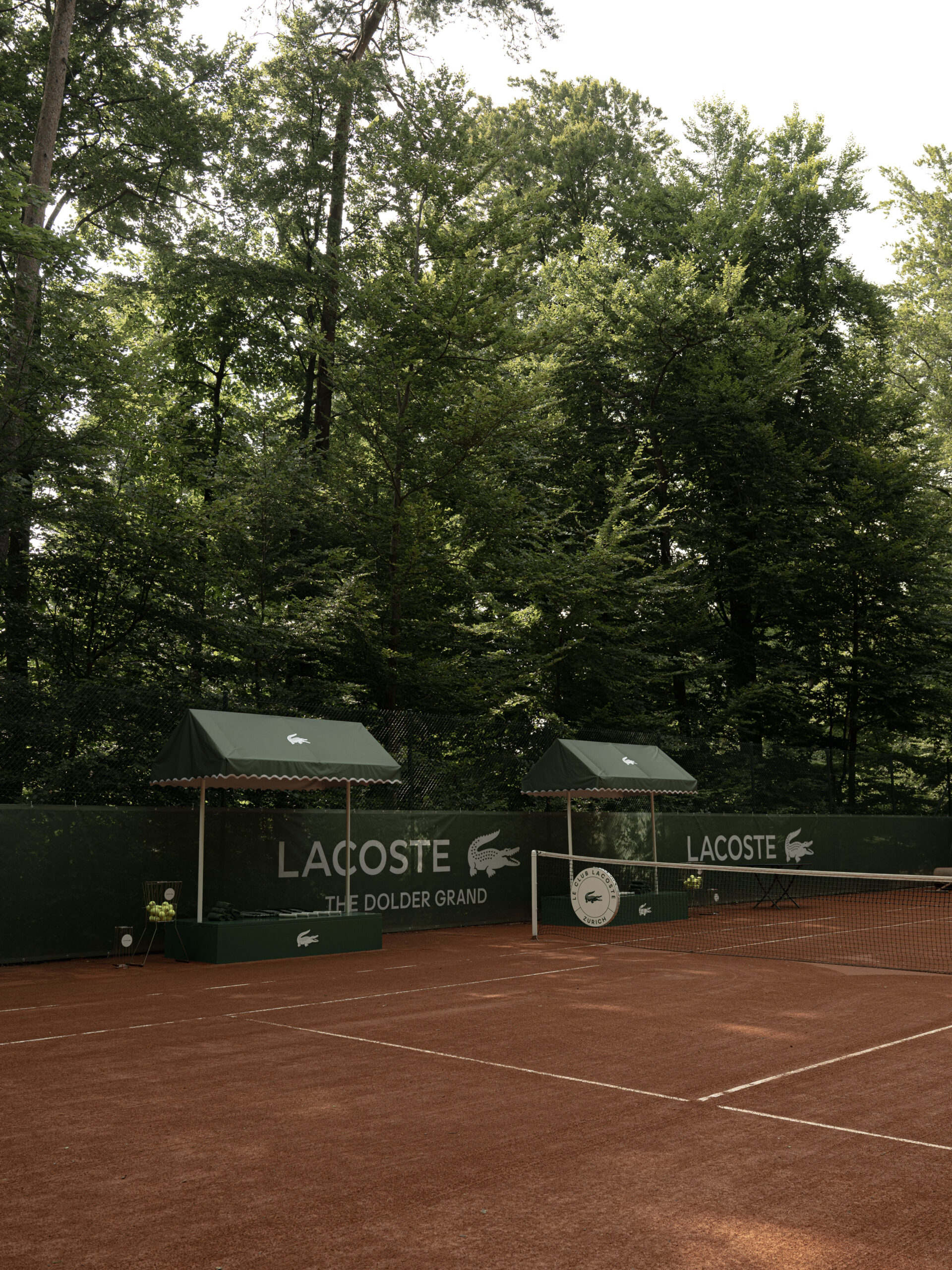 A clay tennis court in Zurich, surrounded by trees and green Lacoste-branded awnings, sits calmly beneath the gaze of the Dolder Grand fairytale hotel—its peaceful scene undisturbed, awaiting its next match.
