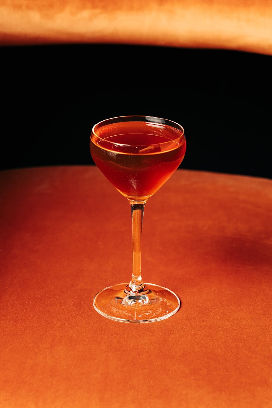 A coupe glass filled with a dark amber cocktail sits on a smooth, orange-brown surface, with a similarly coloured background, creating a warm and elegant atmosphere—just the sort of refined scene you’d discover in an insider’s guide to Edinburgh travel.