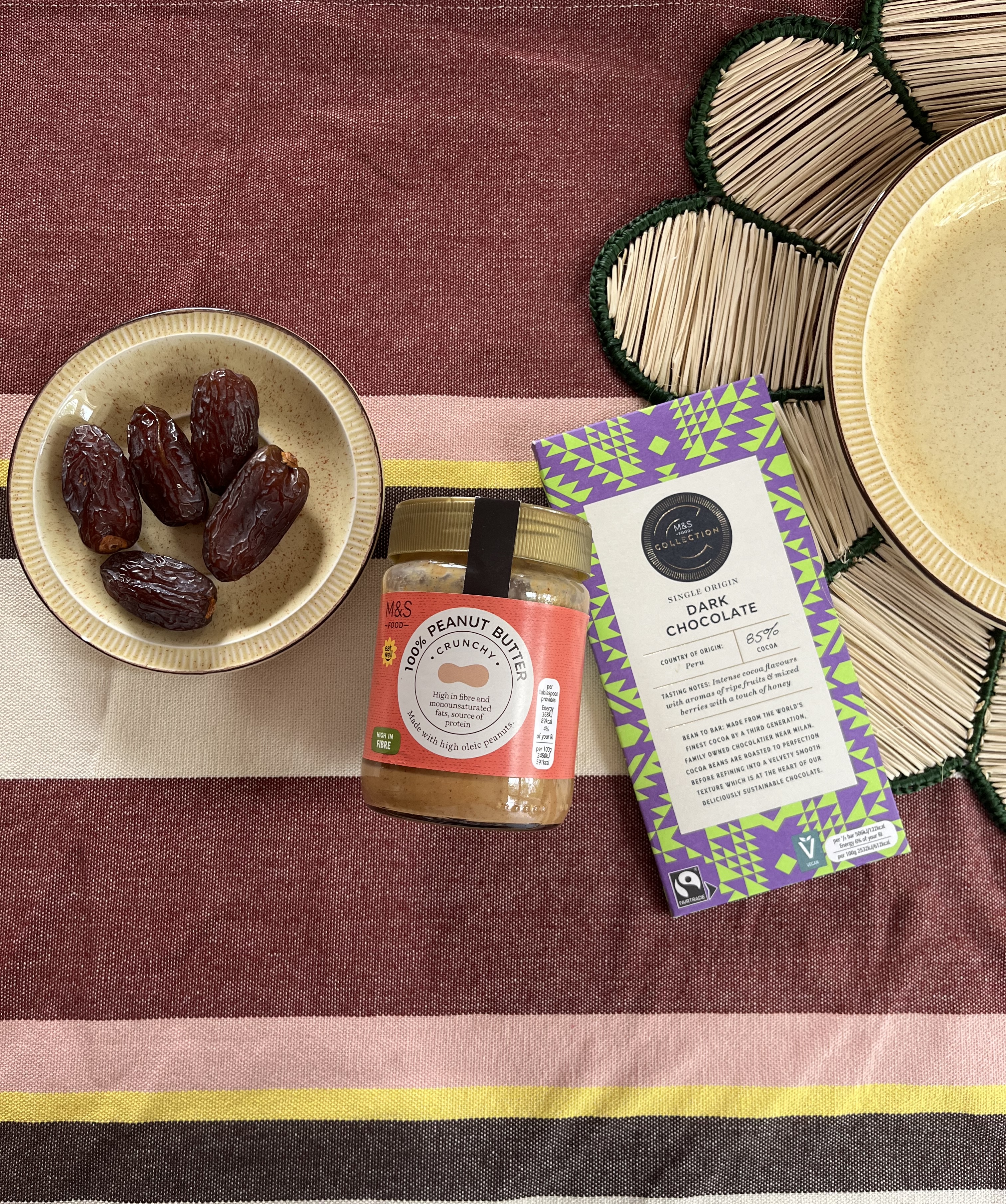 A bowl of peanut butter dates, a jar of crunchy peanut butter, and a bar of dark chocolate sit on a colourful striped tablecloth next to a cream plate with a green-bordered woven placemat.