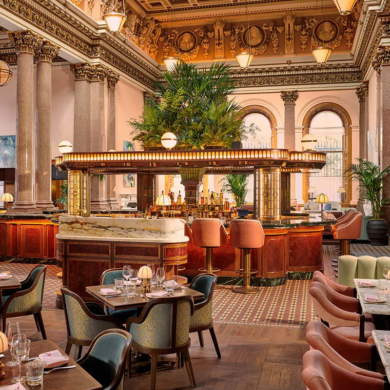 Elegant restaurant with high ornate ceilings, marble columns, and large arched windows—an insider’s guide to one of Edinburgh’s most sophisticated dining experiences. A central bar featuring lush plants and warm lighting is surrounded by plush chairs and set dining tables, making this the perfect spot for travellers seeking refinement and comfort.