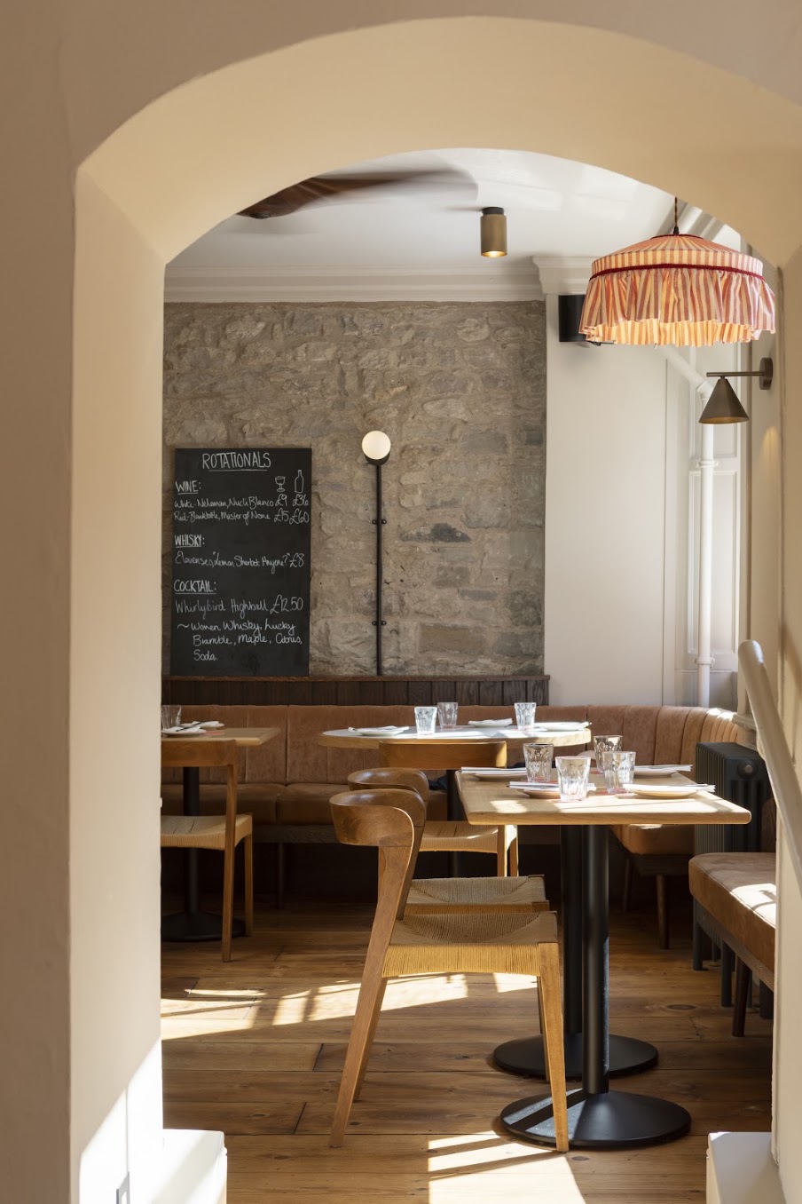 Sunlit restaurant interior with wooden chairs, set tables, a booth, and a chalkboard specials menu on a stone wall—an atmosphere often highlighted in any Insider’s Guide to Edinburgh. A red-striped lampshade hangs from the ceiling near a bright window, adding to the charm of this inviting Edinburgh eatery.