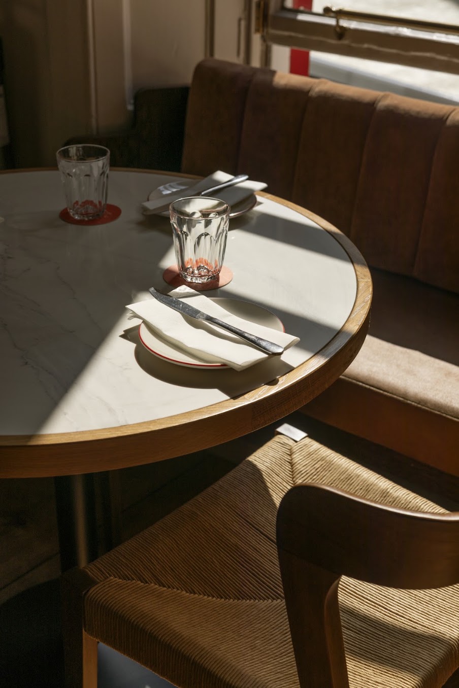 A round table set for two with glasses, serviettes, and cutlery in a sunlit Edinburgh café. A brown upholstered bench and wooden chair are partially visible, evoking the charm of a local spot featured in an insider’s guide to travel.