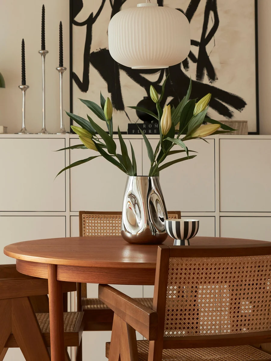 A round wooden dining table with two cane chairs, a statement vase filled with lilies, a striped bowl, black candlesticks, a white pendant lamp, and abstract art in the background.