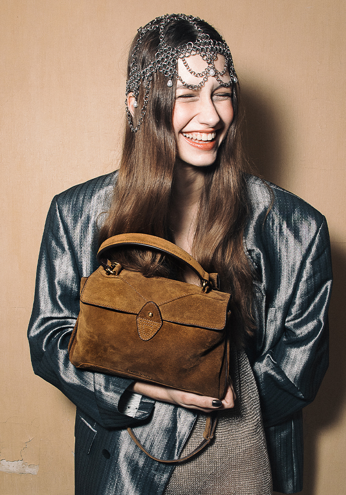 Smiling woman with long brown hair wears a metallic jacket and a beaded headpiece, holding a brown suede handbag from small fashion brands against a beige wall.