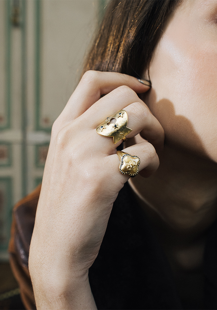 A close-up of a person’s hand wearing two unique gold rings with organic, textured designs from small fashion brands, held near their face. The background is softly blurred, showing part of the person’s brown hair and a decorative wall.