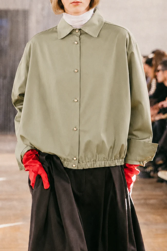 Person wearing a light green buttoned jacket, white polo neck, black skirt, and bright red gloves—an ensemble that could easily come from one of today’s innovative small fashion labels. The background shows blurred people seated, suggesting a fashion show.