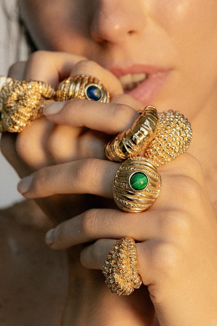 A person holds their hand near their face, displaying multiple ornate gold rings with textured designs and colourful gemstones in green and blue, each thoughtfully crafted by small fashion brands.