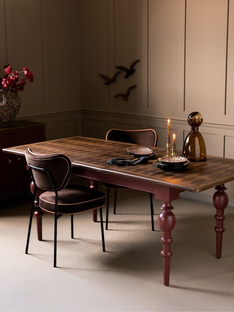 A Rockett St George rectangular wooden dining table with a chequered pattern, set with two lit candles and a glass carafe, surrounded by two upholstered chairs with curved backs.