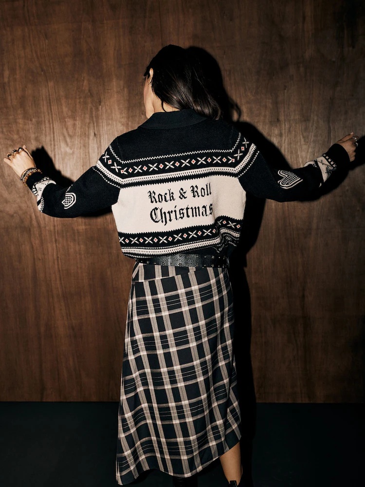 A person seen from behind wears a Rockett St George black and cream patterned jumper with the text Rock & Roll Christmas on the back, paired with a checked midi skirt.